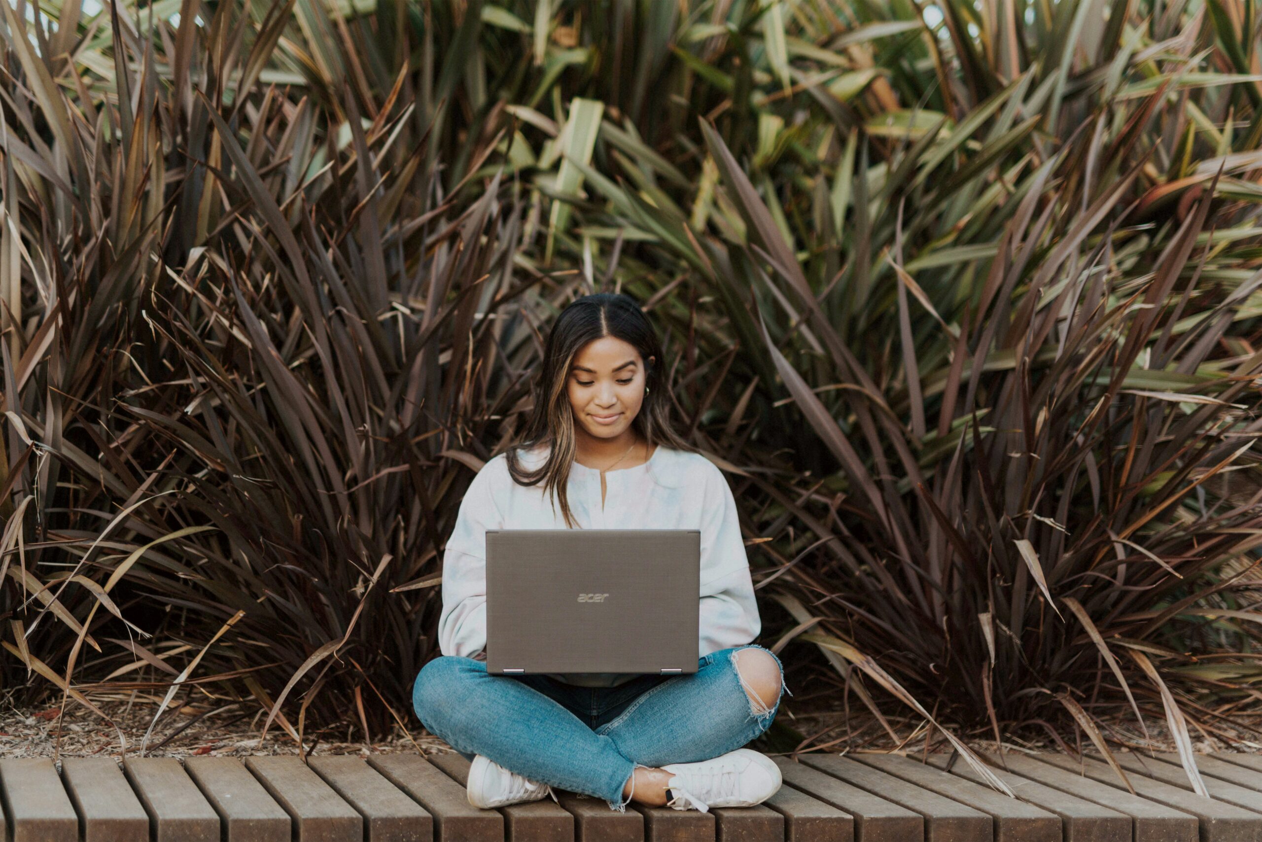 Young woman sitting outdoors with a laptop, working remotely on a wooden platform surrounded by tall plants; representing flexible, remote internship opportunities with Ace Partners.